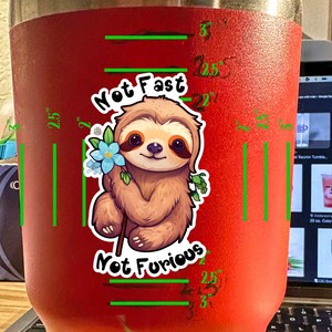 May include: Red stainless steel tumbler with a sloth cartoon graphic and the text "Not Fast Not Furious". The tumbler has a measurement scale in inches.
