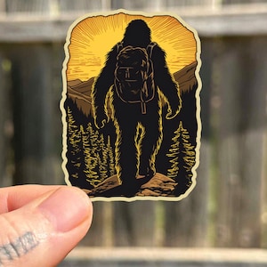 May include: A black and white illustration of a Bigfoot figure wearing a backpack and walking through a forest. The background is a sunset with a yellow sky and orange mountains.