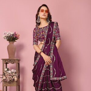 May include: A purple silk saree with silver sequins and floral embroidery. The saree has a scalloped border with pink and white accents. The saree is paired with a matching purple blouse with floral embroidery.