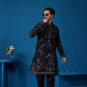 May include: Black tunic with a floral and bird pattern, paired with black trousers. The tunic has a mandarin collar and long sleeves. The outfit is completed with brown leather shoes. The background is a solid blue.