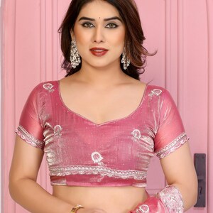 May include: A pink sari blouse with short sleeves and a V-neck. The blouse has white embroidered details and a scalloped trim. The fabric has a subtle sheen. The woman is wearing silver earrings and a gold watch.