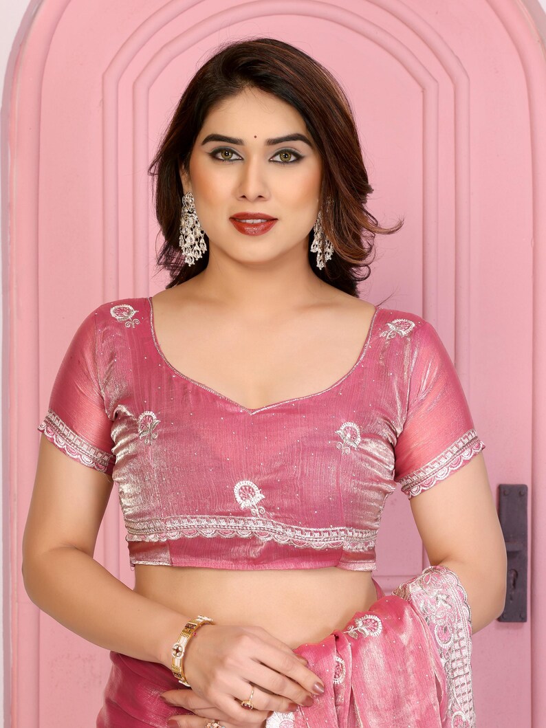 May include: A pink sari blouse with short sleeves and a V-neck. The blouse has white embroidered details and a scalloped trim. The fabric has a subtle sheen. The woman is wearing silver earrings and a gold watch.