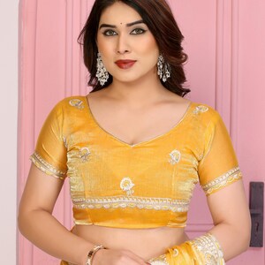 May include: Mustard yellow blouse with short sleeves and a V-neck. The blouse features embroidered detailing along the neckline and sleeves. The fabric appears to be a soft velvet. The blouse is part of a traditional Indian outfit.