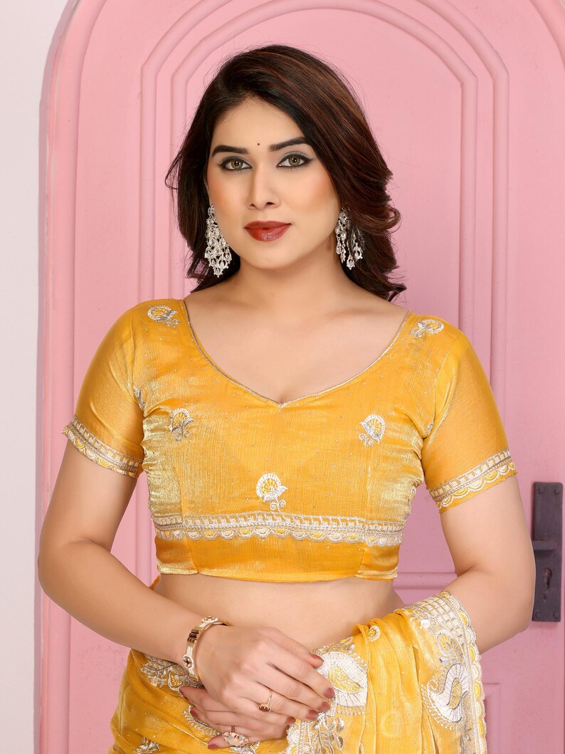 May include: Mustard yellow blouse with short sleeves and a V-neck. The blouse features embroidered detailing along the neckline and sleeves. The fabric appears to be a soft velvet. The blouse is part of a traditional Indian outfit.