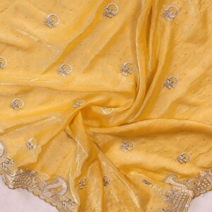May include: A flowing, yellow fabric with intricate embroidery. The fabric features small, repeating floral patterns and a detailed border with a paisley design. The material has a subtle sheen, suggesting a luxurious texture.