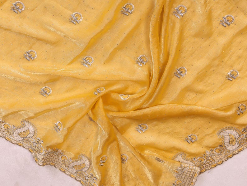 May include: A flowing, yellow fabric with intricate embroidery. The fabric features small, repeating floral patterns and a detailed border with a paisley design. The material has a subtle sheen, suggesting a luxurious texture.