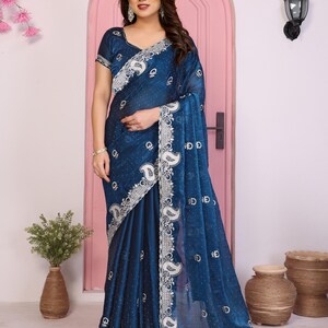 May include: A dark teal saree with intricate silver embroidery along the borders and detailing. The saree features a paisley pattern and a sheer, flowing fabric. The model is wearing the saree in a studio setting.