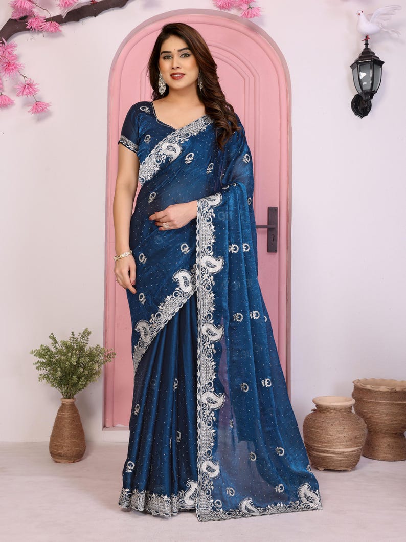 May include: A dark teal saree with intricate silver embroidery along the borders and detailing. The saree features a paisley pattern and a sheer, flowing fabric. The model is wearing the saree in a studio setting.
