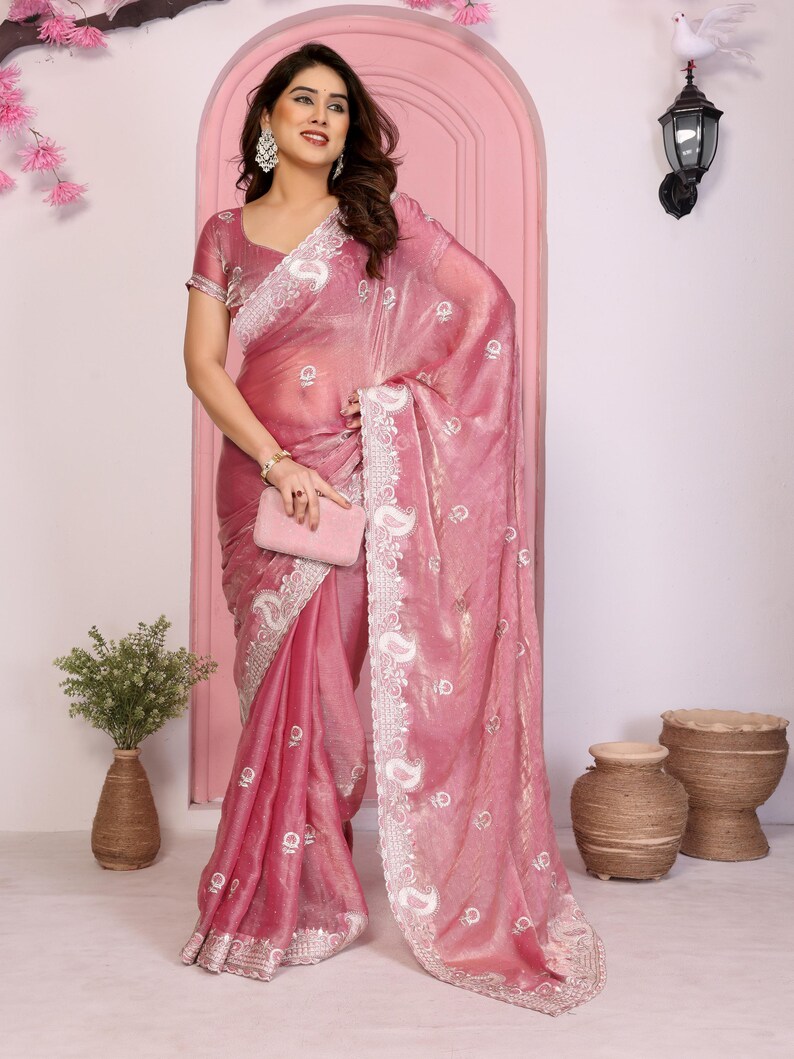 May include: A pink saree with intricate white embroidery and a shimmering, translucent fabric. The saree features a detailed border and is paired with a matching blouse. A small pink clutch is held in the hand. The overall look is elegant and traditional.