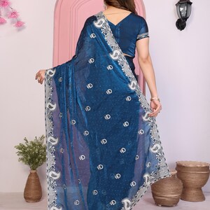 May include: A teal saree with intricate white embroidery along the border and scattered across the fabric. The saree features a detailed paisley design and a matching teal blouse. The fabric has a shimmering, translucent quality.
