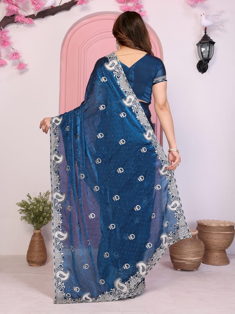 May include: A teal saree with intricate white embroidery along the border and scattered across the fabric. The saree features a detailed paisley design and a matching teal blouse. The fabric has a shimmering, translucent quality.