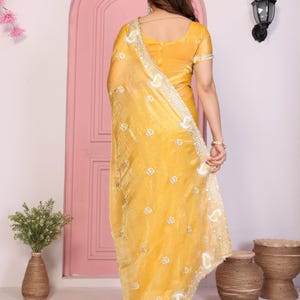 May include: A woman wearing a mustard yellow sari with intricate embroidery and a matching blouse. The sari features a delicate, shimmering fabric with a detailed border. The outfit is accessorized with jewelry.