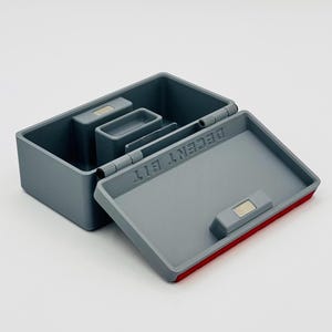 May include: Gray rectangular box with an open lid, revealing interior compartments. The lid has the text "DECENT BIT" printed on it. A red accent lines the edge of the lid. The box has a magnetic closure.