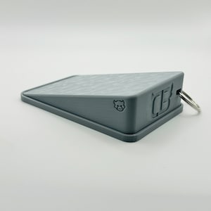 May include: A gray plastic wedge doorstop with a textured surface and a small key ring attached. The doorstop has a small logo of a bull's head and the letter "B" on the side.