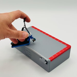 May include: A miniature gray scooter ramp with red trim and a blue and black scooter. The ramp has a rectangular shape with a flat surface. The scooter is held by a hand.