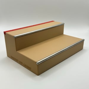 May include: A two-step wooden ramp with metal rails for fingerboarding. The ramp is painted tan with red accents and has the text "DECENT BIT GRIND" printed on the side.