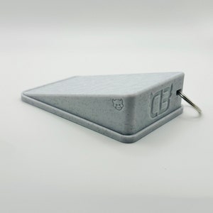 May include: A gray, wedge-shaped doorstop with a small, gray bull logo and the letters "CB" embossed on the side. The doorstop has a metal ring attached to it.