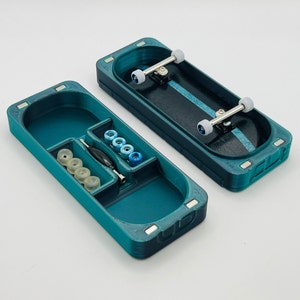 May include: Teal blue fingerboard storage case with a compartment for wheels and a separate compartment for a fingerboard. The case has a fingerboard inside with blue wheels.