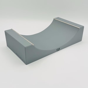 May include: A gray miniature skateboard ramp with a curved surface and metal rails. The ramp has a smooth finish and is designed for fingerboarding. The text "DECKZ BIT" and a logo are visible on the side.