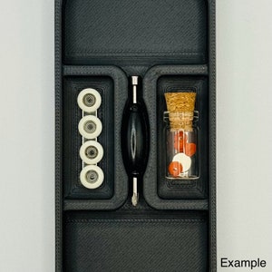 May include: A black plastic storage box with three compartments. The compartments contain a set of five white skateboard wheels, a small bottle with a cork top filled with red and white beads, and a black and silver tool.