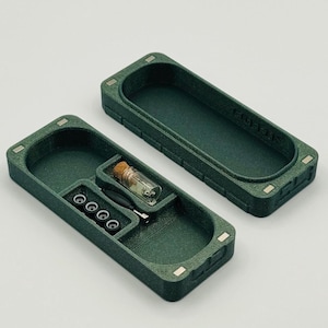 May include: A dark green rectangular storage box with a lid, containing small compartments. Inside are small black objects, a clear glass vial with a cork, and a small tool. The box is open, revealing its contents.