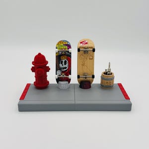 May include: A gray and red display stand with a red fire hydrant, two fingerboards, and a small wooden barrel with a screwdriver inside. The fingerboards are decorated with graphics. One fingerboard has a skull and the other has the text "Slatty Fingers".