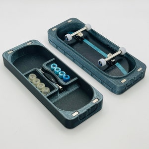 May include: Two-piece fingerboard carrying case and fingerboard. The case is a textured blue-gray with a compartment for the fingerboard and accessories. The fingerboard has a black deck with a blue stripe. Accessories include wheels and a screwdriver.