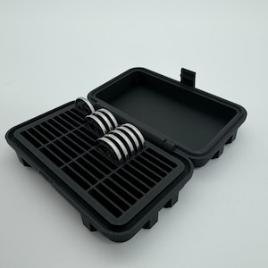 Puede incluir: Una caja de plástico negra con un interior enrejillado para guardar objetos pequeños, como baterías o monedas. La caja está abierta y tiene una fila de objetos redondos de color plateado en su interior.