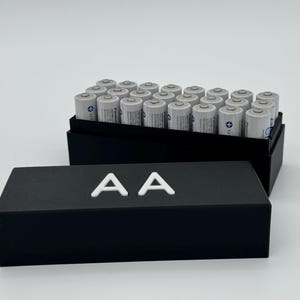 Op de afbeelding: Een zwarte doos met het label "AA" aan de voorkant. De doos is open en bevat een grote hoeveelheid AA-batterijen.