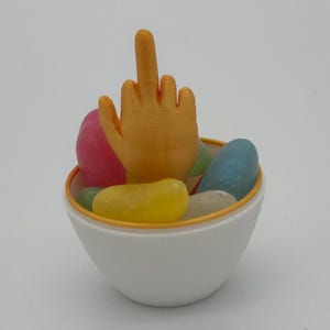 Könnte beinhalten: Eine weiße Schüssel mit goldfarbenem Rand, gefüllt mit bunten Gummibärchen. Ein goldfarbener Mittelfinger ragt aus der Schüssel heraus.