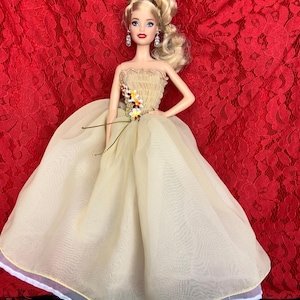 May include: A blonde doll wearing a beige gown with a floral embellishment and a white underskirt. The doll is posed with one hand on her hip and the other holding the gown. The background is a red lace fabric.