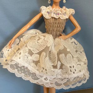 May include: A doll wearing a beige and white lace dress with a ruffled collar and yellow boots. The dress features a fitted bodice and a full skirt with floral lace overlay. The doll has blonde hair styled in an updo and is wearing dangling earrings.