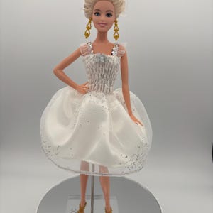 May include: A blonde doll wearing a white dress with silver glitter accents. The dress has a ruched bodice and a full skirt. The doll is wearing gold earrings and gold heels. The doll is standing on a clear stand.