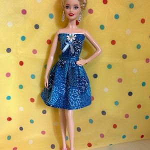 May include: A doll wearing a blue strapless dress with a floral pattern and a white bow. The doll has blonde hair, dangling earrings, and pink heels. The background is yellow with colorful polka dots.