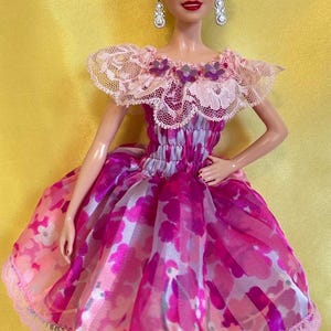 May include: A doll wearing a pink and white floral print dress with a lace trim. The doll has blonde hair and is wearing dangling earrings. The dress has a lace neckline with purple flower accents.