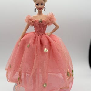 May include: A doll wearing a pink gown with a tulle skirt and gold accents. The doll has blonde hair styled in an updo, dangling earrings, and a floral embellishment on the bodice. The dress features gold sequins and floral appliqués.