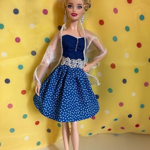 May include: A doll wearing a blue polka dot dress with a white lace belt and a sheer white shawl. The doll has blonde hair styled in an updo, dangling earrings, and pink heels. The background is yellow with colorful polka dots.