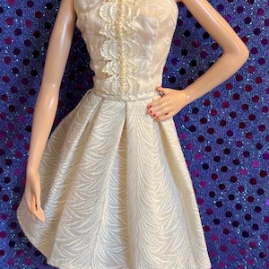 May include: A doll wearing a cream-colored strapless dress with a textured pattern and a pleated skirt. The doll has blonde hair, red lipstick, and pink high heels. She is wearing dangling earrings.