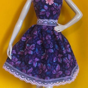 May include: A doll wearing a strapless dress with a purple leaf pattern, a pink lace trim, and matching pink shoes. The doll has blonde hair, dangling earrings, and red lipstick. The background is yellow.