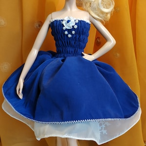 May include: A blonde doll wearing a blue dress with white trim and a white flower detail. The doll is wearing red shoes and has a white background.