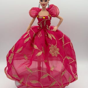 May include: A doll wearing a vibrant pink gown with gold embroidered floral patterns. The dress features puffed sleeves and a full skirt. The doll has blonde hair styled in an updo and is wearing dangling earrings. The gown is embellished with gold trim.