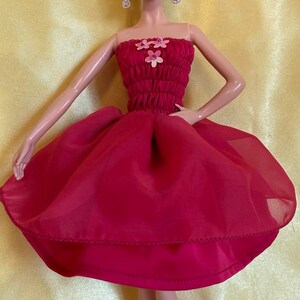 May include: A doll wearing a red strapless dress with a full skirt and flower accents. The doll has blonde hair, dangling earrings, and red cowboy boots. The background is a solid yellow.