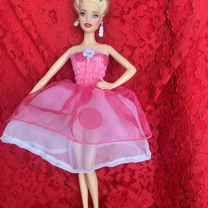 May include: A blonde doll wearing a pink and white dress with a flower detail on the bodice. The doll is standing on a red lace background.