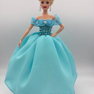 May include: A fashion doll wearing a light blue gown with a full skirt and off-the-shoulder sleeves. The doll has blonde hair styled in an updo, dangling earrings, and red shoes. The bodice is embellished with jewels.