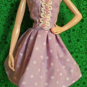 May include: A doll wearing a lavender dress with white polka dots and a white lace bodice with pink accents. The doll has blonde hair styled in a bun and is wearing red shoes.