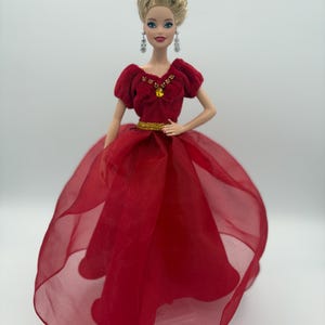 May include: A doll wearing a red gown with a sheer skirt. The doll has blonde hair styled in an updo and is wearing dangling earrings. The bodice of the dress is velvet and adorned with a gold-colored belt and a yellow jewel.