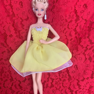 May include: A blonde doll wearing a yellow dress with a white lace trim. The doll has blue eyes and is wearing earrings. The doll is standing on a red lace background.