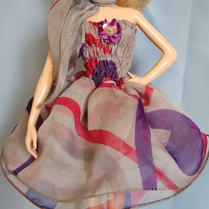 May include: A fashion doll wearing a gray dress with red and purple accents. The dress has a sheer overlay with a plaid pattern. The doll has blonde hair and is wearing red heels. The dress is adorned with a floral embellishment.