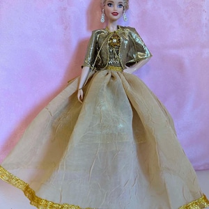 May include: A doll wearing a gold-colored gown with a matching bolero jacket. The doll has blonde hair and is wearing dangling earrings. The gown has a full skirt with gold trim. The doll is posed against a pink background.