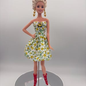 May include: A doll wearing a strapless white dress with a yellow and green floral pattern. The doll has blonde hair, gold earrings, and red cowboy boots. A yellow and blue brooch accents the dress.